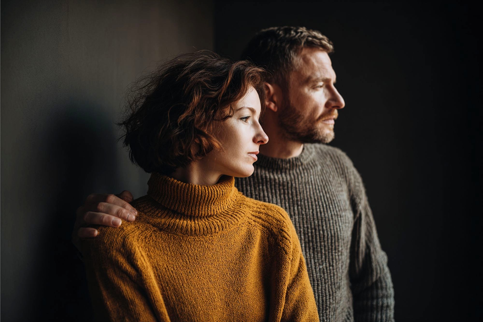 A woman in a mustard-colored sweater and a man in a gray sweater stand close together, looking thoughtfully out of frame toward the light. The man's arm rests gently on the woman's shoulder.