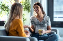 Two women sit on couches in a bright living room, having a friendly conversation. One woman, smiling and holding a yellow mug, gestures with her hand while the other listens attentively.