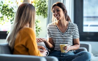 Two women sit on couches in a bright living room, having a friendly conversation. One woman, smiling and holding a yellow mug, gestures with her hand while the other listens attentively.