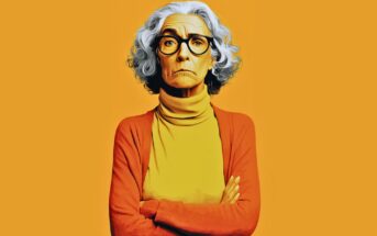 An older woman with gray curly hair, wearing glasses, a yellow turtleneck, and an orange cardigan, stands with arms crossed against a solid orange background, looking serious.