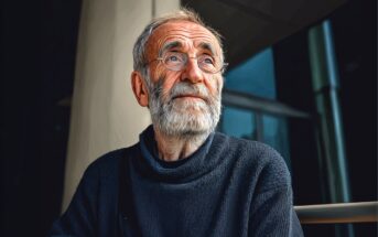An elderly man with glasses and a white beard, wearing a dark sweater, sits indoors and looks thoughtfully into the distance, illuminated by soft light from a window.