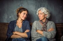 A young woman and an older woman sit side by side on a couch with their arms crossed; the young woman is smiling while the older woman looks serious. Both face each other against a dark, textured wall.