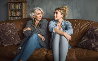 An older woman with gray hair and a younger woman with blonde hair sit on a brown leather couch, facing each other, arms crossed, and making eye contact in a cozy living room setting.