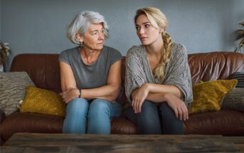 Two women sit on a brown couch, facing each other. The older woman looks concerned, while the younger woman looks away, appearing upset. Both have light hair and casual clothes. Cozy cushions and plants are in the background.