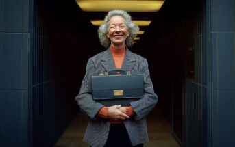An older woman with gray hair smiles warmly while holding a briefcase. She wears a pinstripe blazer over a red turtleneck and stands in a hallway with overhead lights and dark walls.