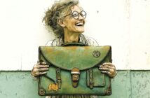 An older woman with curly hair and round glasses smiles while holding a large, worn green satchel in front of her, standing against a two-toned wall.