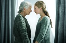 An elderly woman and a young woman stand face to face, gazing intently at each other. Both wear textured green dresses, and soft light streams in from behind, highlighting their expressions and features.