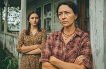 Two women stand on a porch with arms crossed, looking serious. The older woman is in the foreground wearing a plaid shirt; the younger woman is behind her, wearing a floral dress. The scene appears tense and somber.