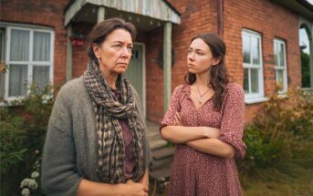 Two women stand outside a brick house, arms crossed, looking at each other with serious expressions. One wears a gray scarf and sweater, the other a red dress. The scene suggests tension or disagreement.