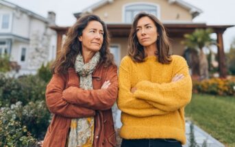 Two women standing with their arms crossed in front of a house, looking in opposite directions.
