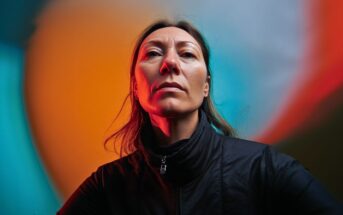 A woman in a dark jacket looks confidently at the camera, lit by dramatic orange and blue lighting with a vibrant, abstract background.
