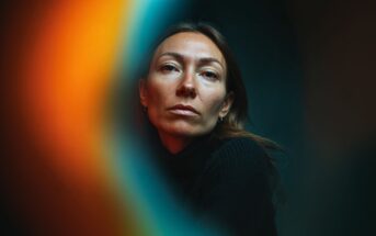 A woman with long brown hair and a serious expression looks at the camera, framed by dark background and vibrant orange and blue blurred light effects in the foreground.