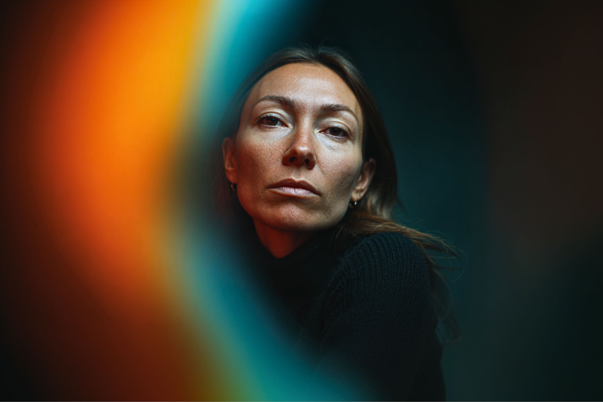 A woman with long brown hair and a serious expression looks at the camera, framed by dark background and vibrant orange and blue blurred light effects in the foreground.