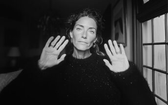 A woman with wavy hair and light-colored eyes sits indoors, facing the camera with both hands raised and palms open. She wears a dark, textured sweater and hoop earrings. Soft light comes through a window on the right.