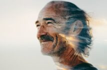 A smiling older man with gray hair and a beard, overlaid with a double exposure of a scenic ocean sunset, blending his profile with waves and sky.