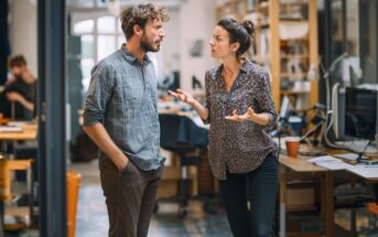 A man and a woman are standing in an office, having a serious conversation. The woman gestures with her hands while the man listens with his hands in his pockets. Desks, computers, and shelves fill the background.