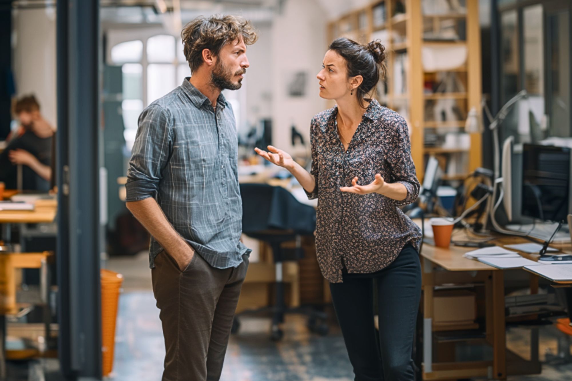 A man and a woman are standing in an office, having a serious conversation. The woman gestures with her hands while the man listens with his hands in his pockets. Desks, computers, and shelves fill the background.
