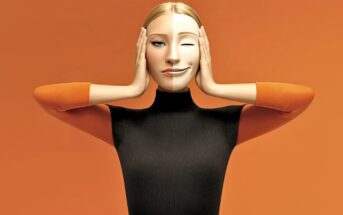 A woman in a black and orange outfit holds a mask over her face. The mask, split down the middle, shows a neutral expression on one side and a winking, smiling face on the other. The background is orange.