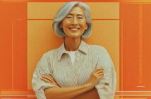A smiling older woman with gray hair and a striped shirt stands with her arms crossed in front of an orange and yellow geometric background.