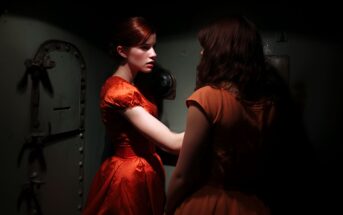 Two women in orange dresses stand closely in a dimly lit room. One faces forward with a serious expression, illuminated by a beam of light, while the other faces away, partially in shadow. An old metal door is in the background.