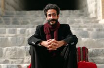 A man with dark hair and a mustache sits on stone steps, smiling. He wears a black coat, a dark red scarf, and has his hands clasped in front of him. A red patterned textile lies beside him on the steps.