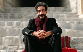 A man with dark hair and a mustache sits on stone steps, smiling. He wears a black coat, a dark red scarf, and has his hands clasped in front of him. A red patterned textile lies beside him on the steps.