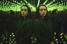 A woman with pale skin and dark hair stands next to a mirrored wall in a room filled with green lights, creating infinite reflections. She wears a dark coat and looks directly at the camera with a serious expression.
