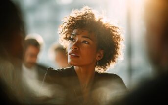 A young woman with curly hair and hoop earrings sits in soft sunlight, looking thoughtful and slightly upward. The background is blurred with other people faintly visible.