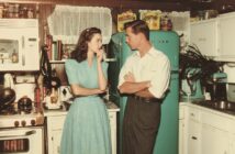 A woman in a blue dress and a man in a white shirt talk in a vintage kitchen with white cabinets and a turquoise fridge, surrounded by retro appliances and food packages.
