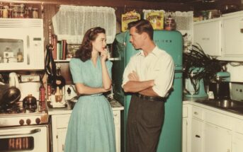 A woman in a blue dress and a man in a white shirt talk in a vintage kitchen with white cabinets and a turquoise fridge, surrounded by retro appliances and food packages.