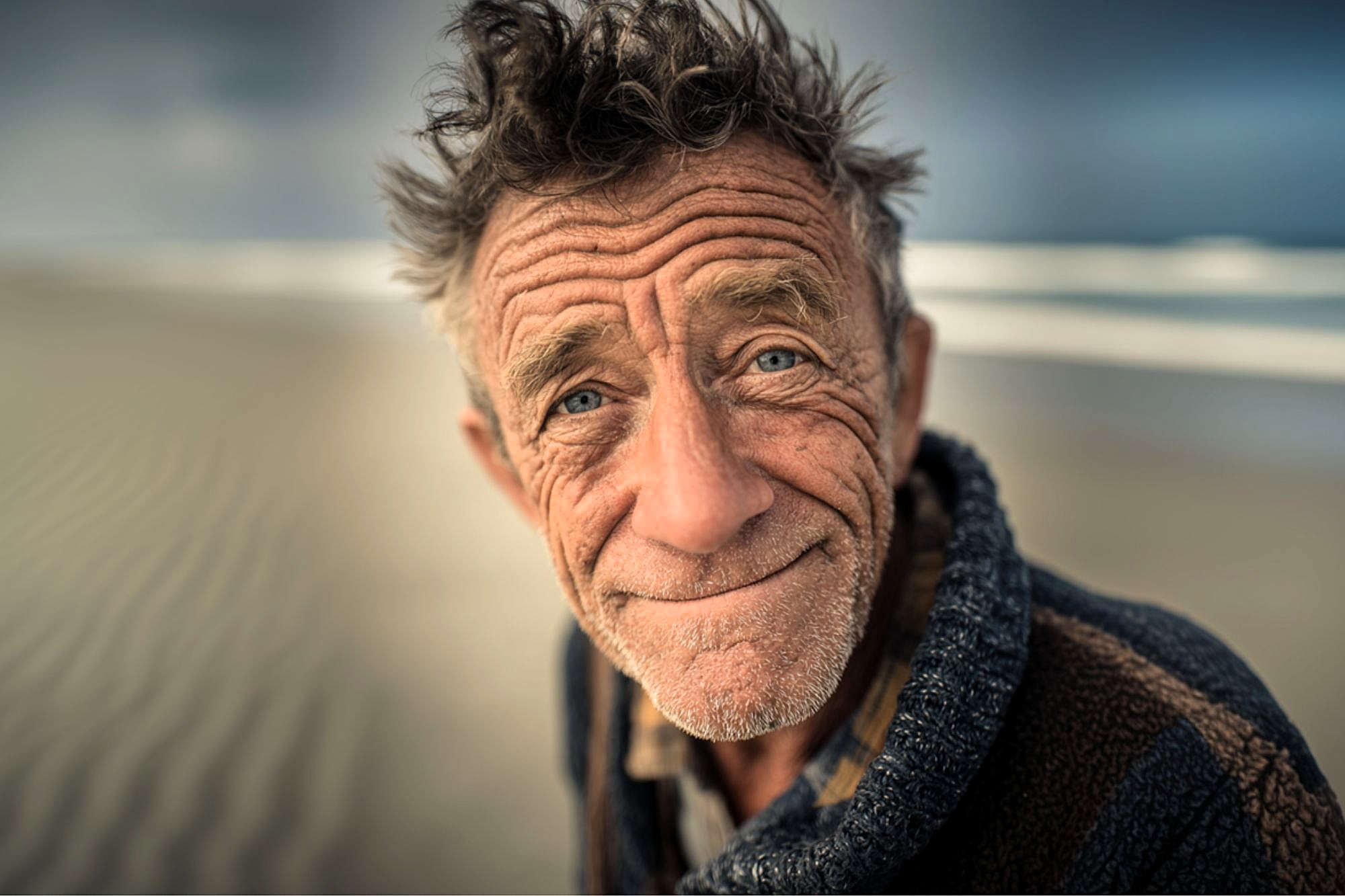 An older man with tousled hair and deep wrinkles smiles gently at the camera, standing on a sandy beach with blurred waves and sky in the background. He wears a cozy, textured sweater.