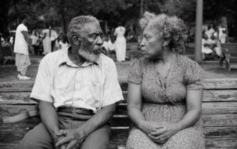 An elderly man and woman sit closely on a worn wooden bench in a park, facing each other and appearing deep in conversation. People and trees are visible in the blurred background. The image is in black and white.