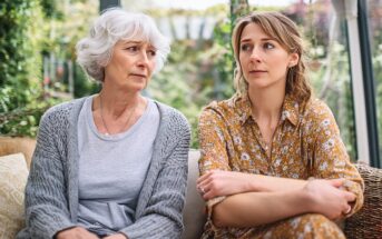 An older woman with gray hair and a younger woman with light brown hair sit next to each other indoors, both looking concerned and thoughtful. The younger woman has her arms crossed, and greenery is visible outside the windows.