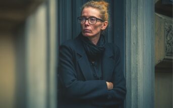A woman with glasses and a black coat stands against a building doorway, arms crossed, looking to the side with a serious expression. The background shows architectural details in muted colors.