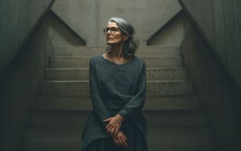 An older woman with gray hair and glasses sits on concrete stairs, hands clasped in her lap, wearing a dark sweater. She gazes thoughtfully to the side in a softly lit, minimalist setting.
