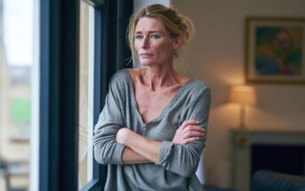 A woman with blonde hair stands indoors by a window, arms folded, gazing outside with a thoughtful and serious expression. She wears a loose grey top and natural light illuminates her face.