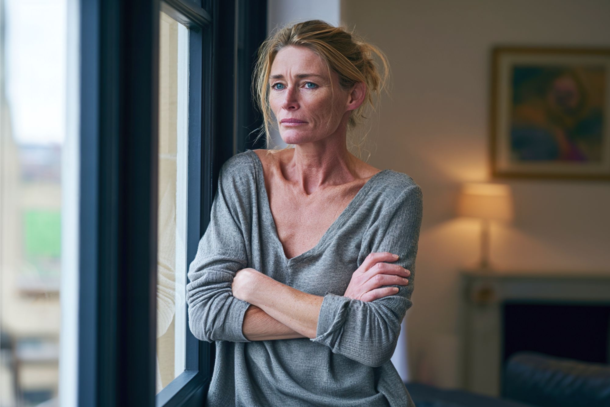 A woman with blonde hair stands indoors by a window, arms folded, gazing outside with a thoughtful and serious expression. She wears a loose grey top and natural light illuminates her face.