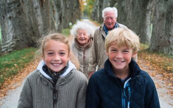 Two smiling children stand in front of an elderly man and woman on a tree-lined path in autumn, with fallen leaves on the ground and tall trees in the background.