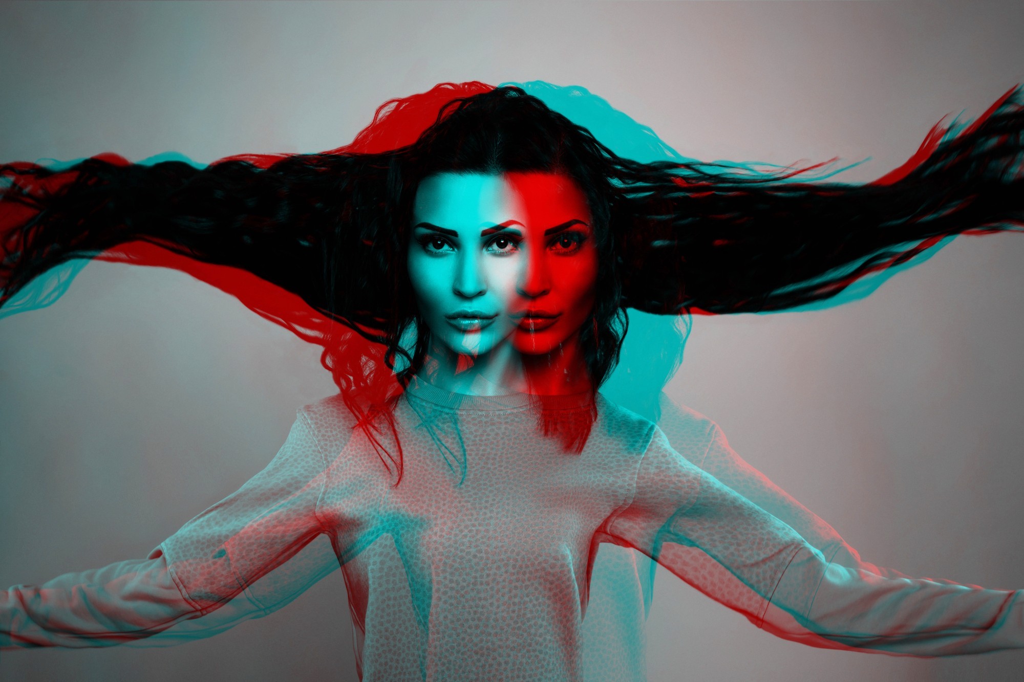 A woman with long, flowing hair is shown with a double exposure and red-blue 3D effect, creating two overlapping images of her face and outstretched arms against a plain background.