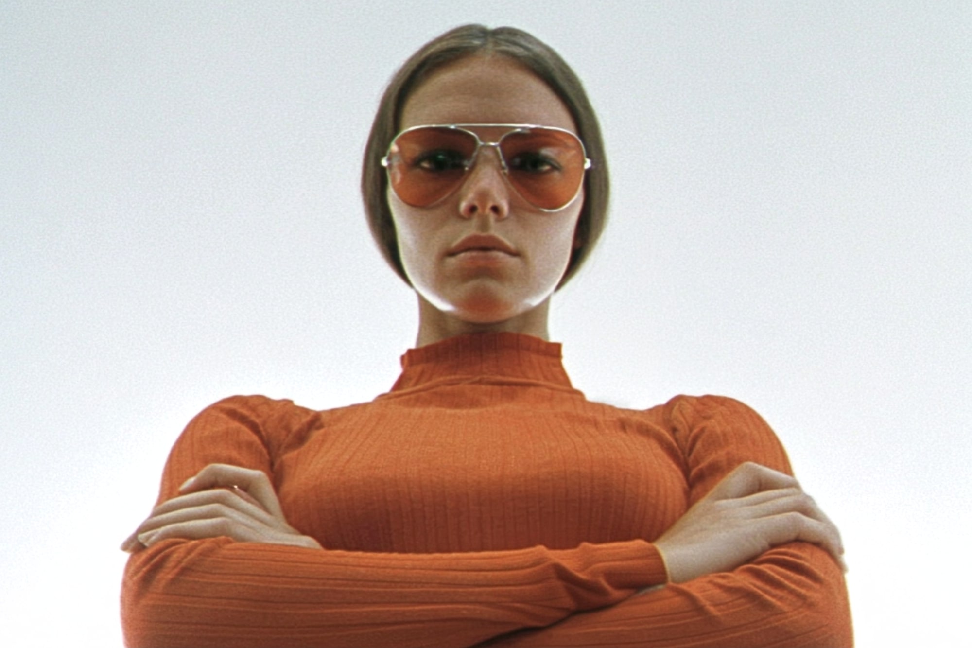 A woman in an orange turtleneck and large tinted sunglasses stands with arms crossed, looking down with a serious expression against a plain light background.