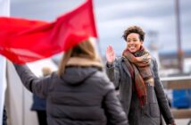 A smiling woman in a coat and scarf waves at someone holding a red flag outdoors on a cloudy day.