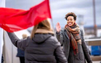 A smiling woman in a coat and scarf waves at someone holding a red flag outdoors on a cloudy day.
