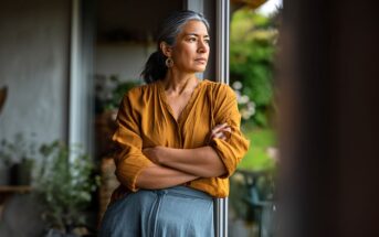 A woman with gray hair in a ponytail, wearing a mustard-yellow blouse and blue skirt, stands indoors by a window with her arms crossed, gazing thoughtfully outside. Plants and greenery are visible in the background.