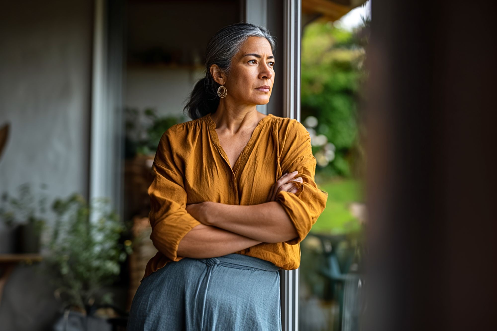 A woman with gray hair in a ponytail, wearing a mustard-yellow blouse and blue skirt, stands indoors by a window with her arms crossed, gazing thoughtfully outside. Plants and greenery are visible in the background.