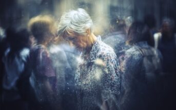 An elderly woman with short gray hair looks down, surrounded by blurred, indistinct figures in a busy, dreamy urban setting, conveying a sense of loneliness or introspection amid a crowd.