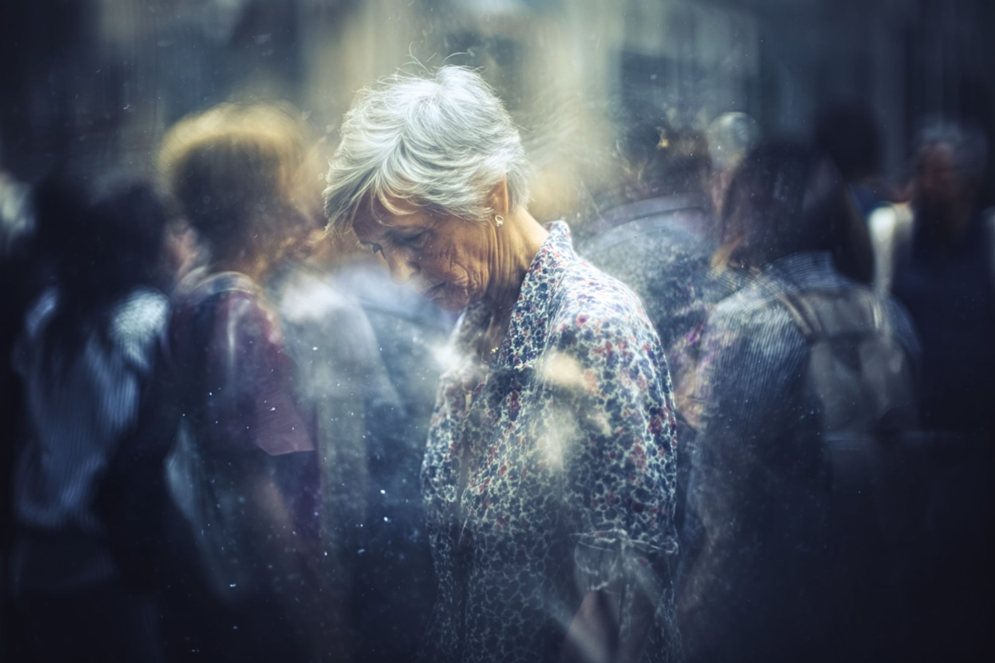 An elderly woman with short gray hair looks down, surrounded by blurred, indistinct figures in a busy, dreamy urban setting, conveying a sense of loneliness or introspection amid a crowd.