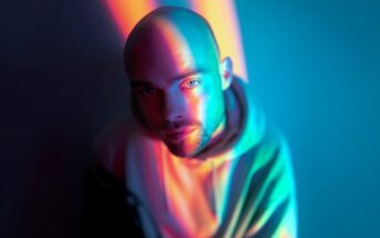 A man in a hoodie with a shaved head is illuminated by vibrant blue, pink, and orange lights, casting colorful shadows on his face and background in a dramatic, artistic portrait.