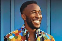 A smiling man with short hair and a beard stands in front of a blue wall, wearing a colorful, patterned shirt with hexagonal designs.