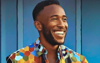 A smiling man with short hair and a beard stands in front of a blue wall, wearing a colorful, patterned shirt with hexagonal designs.