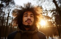 A man with curly hair and a beard stands outdoors in a winter forest at sunset, wearing a jacket. Sunlight shines through the trees behind him, creating a warm, glowing effect around his hair.